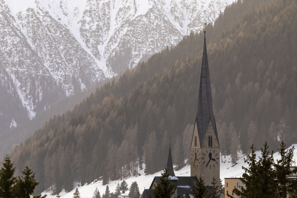 A church with a tall spire located in a mountainous area, surrounded by snow-covered trees and slopes.
