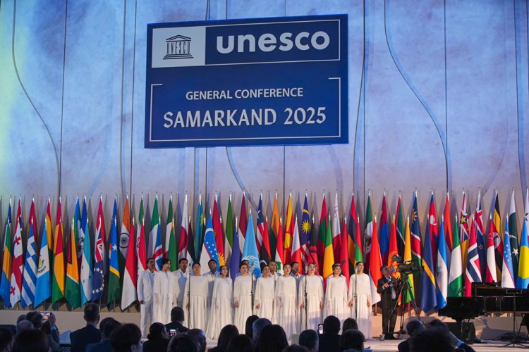 A choir of performers in white outfits singing on stage at the UNESCO General Conference in Samarkand 2025, with a backdrop of flags from various countries and a large UNESCO banner.