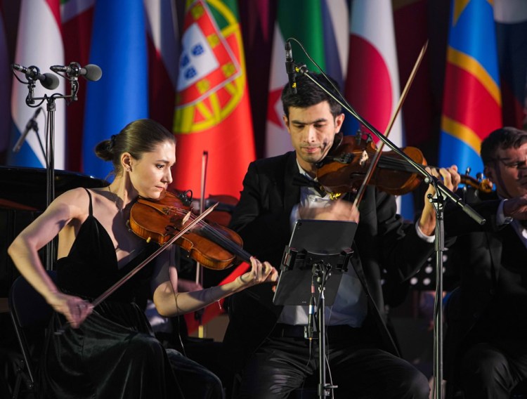 A female and a male violinist performing together on stage, with international flags displayed in the background.