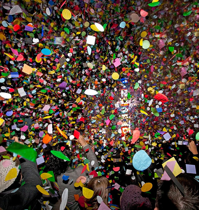 A colorful shower of confetti falls over a crowd celebrating New Year's Eve in Times Square, with excited hands reaching up to catch the vibrant pieces.