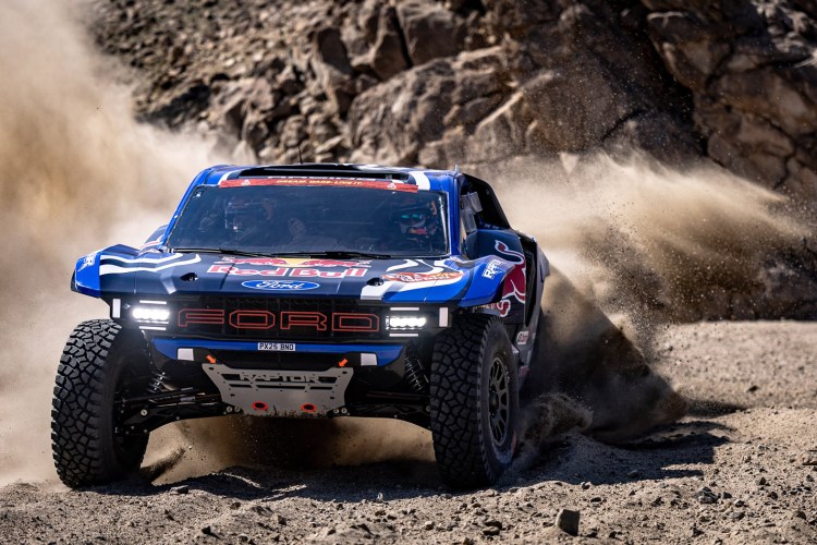 A Ford off-road truck racing on a sandy terrain, kicking up dust and debris, with rocky terrain in the background. The vehicle features a blue and red design with sponsorship logos.