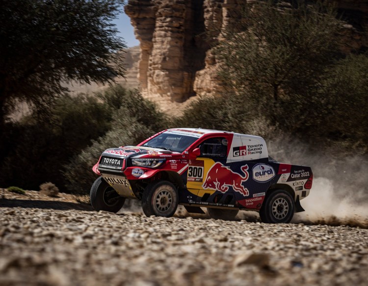 A red and white racing truck driving on a rocky terrain, kicking up dust in a desert landscape with rocky formations in the background.
