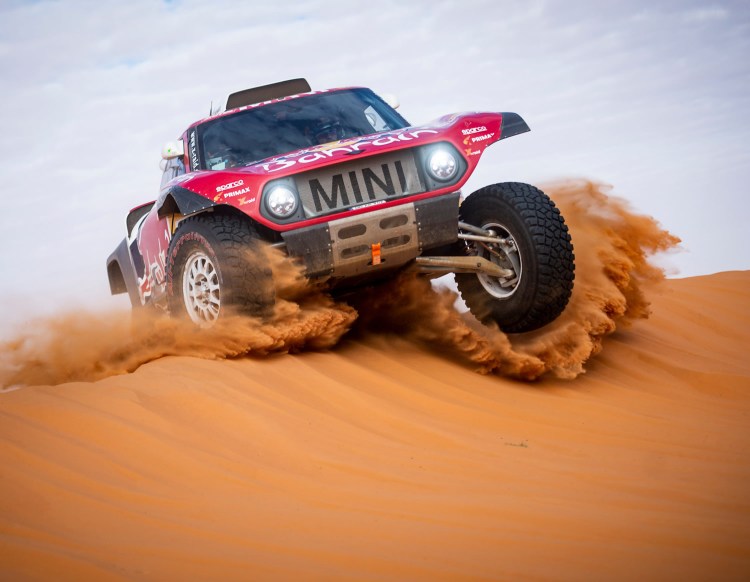 A red off-road race car, labeled 'MINI', speeds across a sandy dune, kicking up dust and sand.