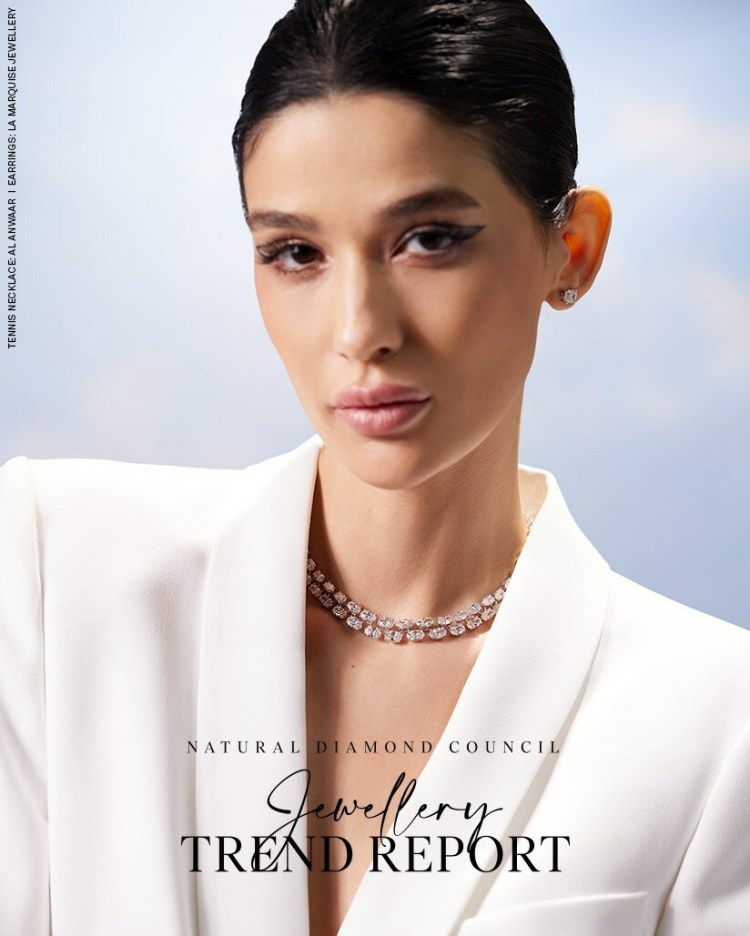 Close-up portrait of a woman wearing a white blazer and elegant diamond necklace, with a soft blurred background.
