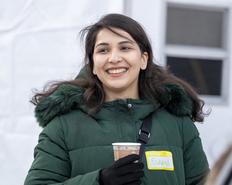 A young woman wearing a green puffer jacket and holding a coffee cup, smiling while standing indoors. She has a name tag that reads 'Hello, my name is Sidhi'.