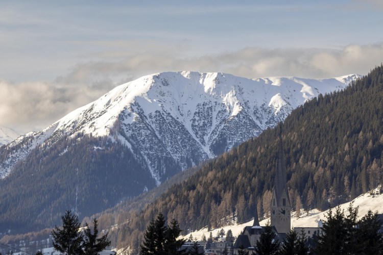 Snow-covered mountains with a church steeple in the foreground, surrounded by evergreen trees and a clear sky.