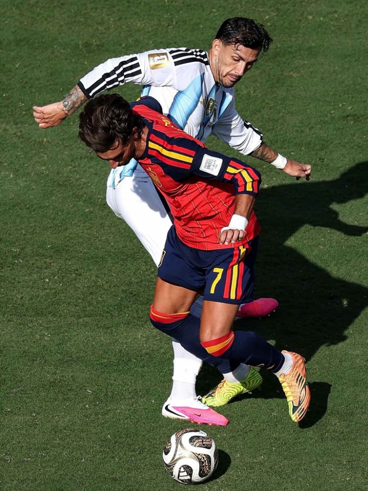 A soccer match scene showing two players in action, one in a red jersey with the number 7 and the other in a white jersey. The player in the red jersey is maneuvering the ball while being challenged from behind by the player in white.