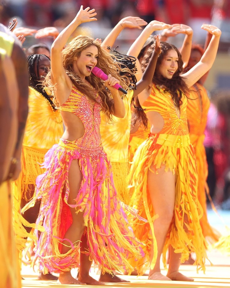 Two performers dancing on stage, wearing colorful yellow and pink costumes. One performer holds a microphone and sings energetically while the other joins in the dance.