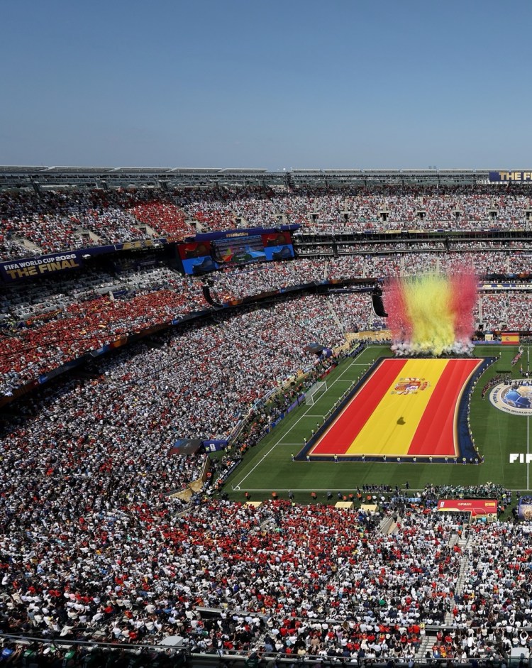 Aerial view of a packed stadium during the FIFA World Cup 2026 final, featuring a large Spanish flag on the field and colorful fireworks in the background.