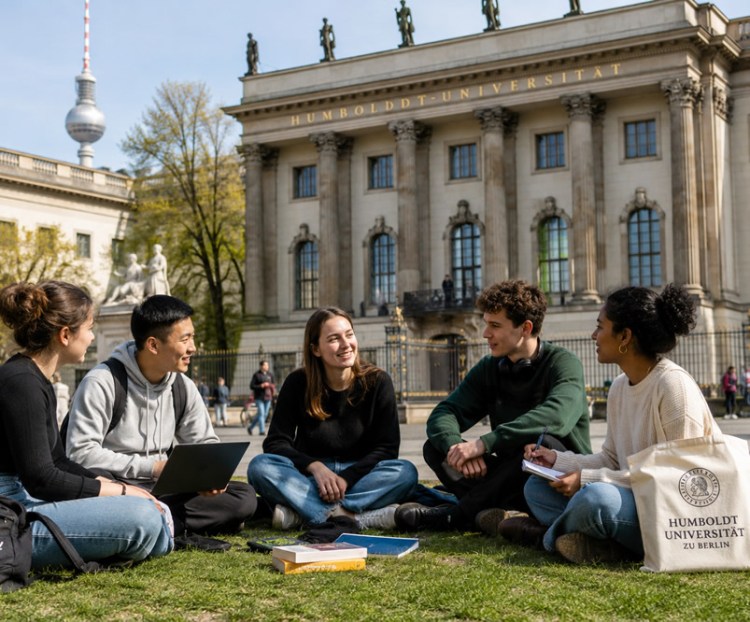 Five university students sitting on the grass in front of Humboldt University in Berlin, engaging in a discussion with a laptop and books nearby.
