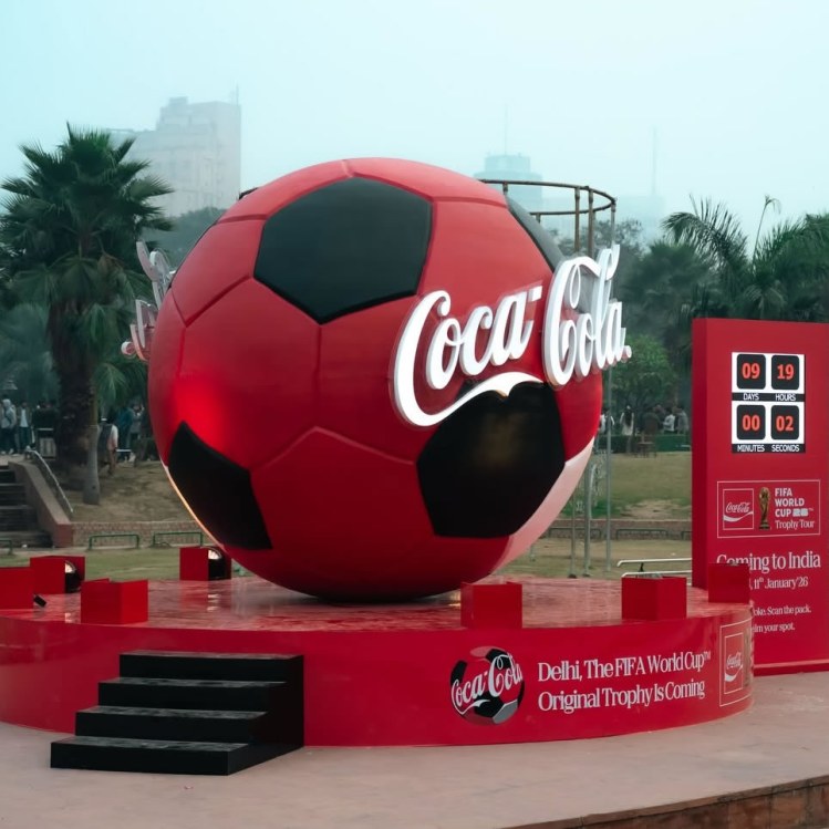 A large Coca-Cola branded soccer ball sculpture displayed outdoors, with a countdown timer indicating 9 days, 19 hours, and 2 seconds until an event, set against a backdrop of palm trees and city buildings.
