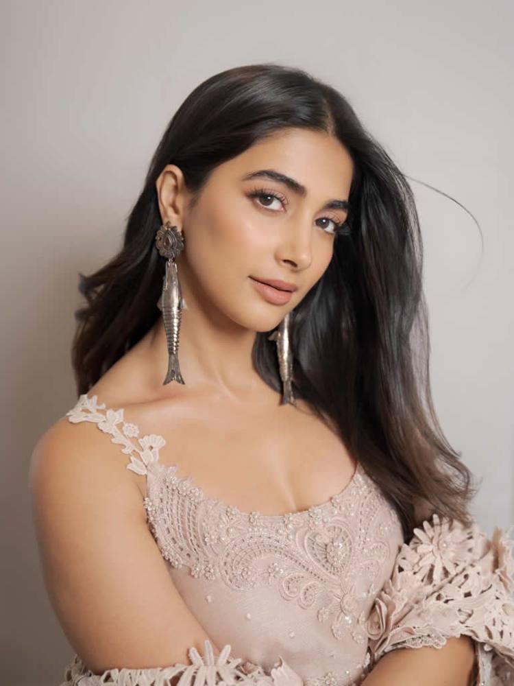 A close-up portrait of a woman with long, dark hair, wearing large, decorative earrings and a pale pink embroidered dress. She poses with a gentle smile against a neutral background.